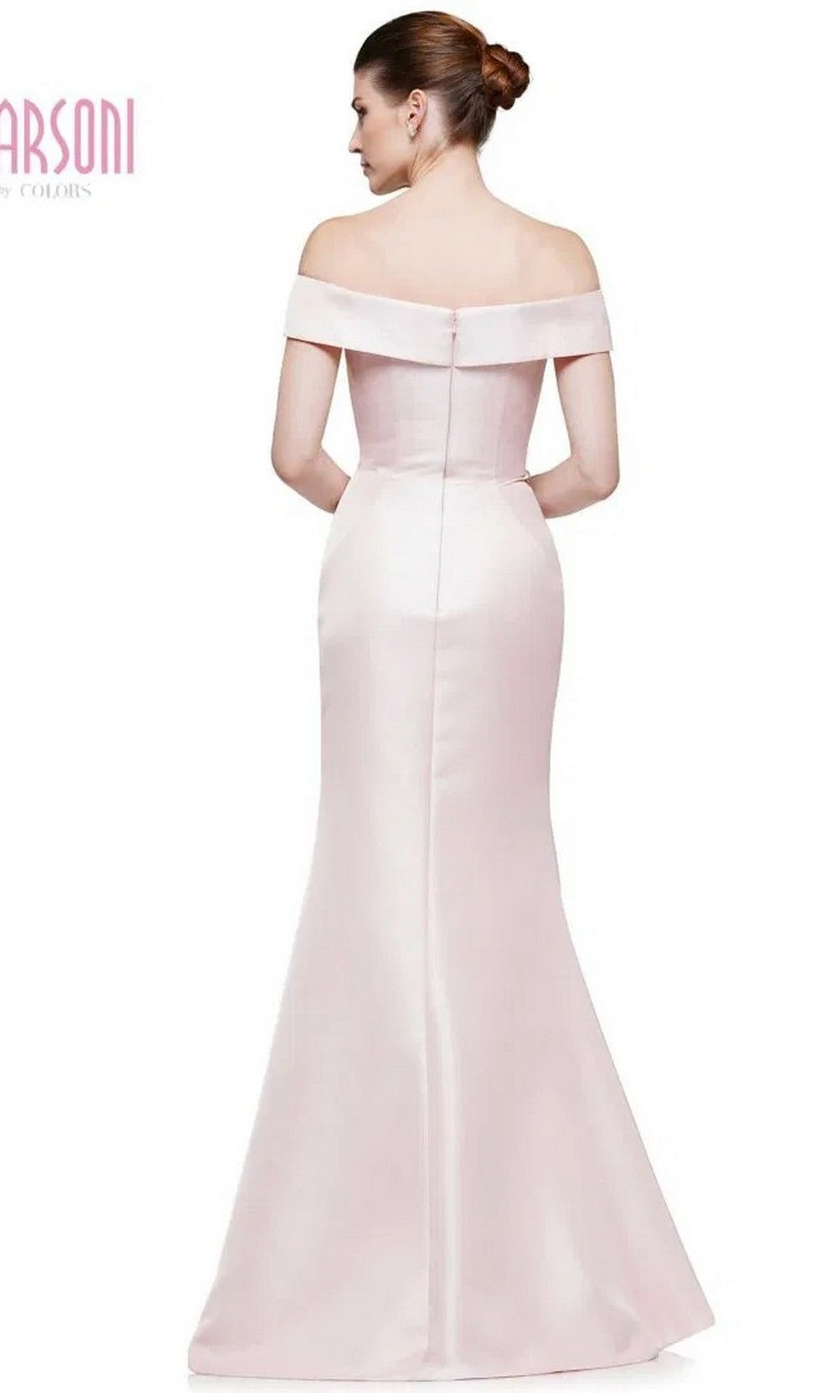 A woman stands facing away, wearing the Marsoni MV1003 long formal dress—an off-the-shoulder satin gown in pale pink with a fitted bodice and flared skirt. Her elegant look is completed with her hair styled in a neat bun.